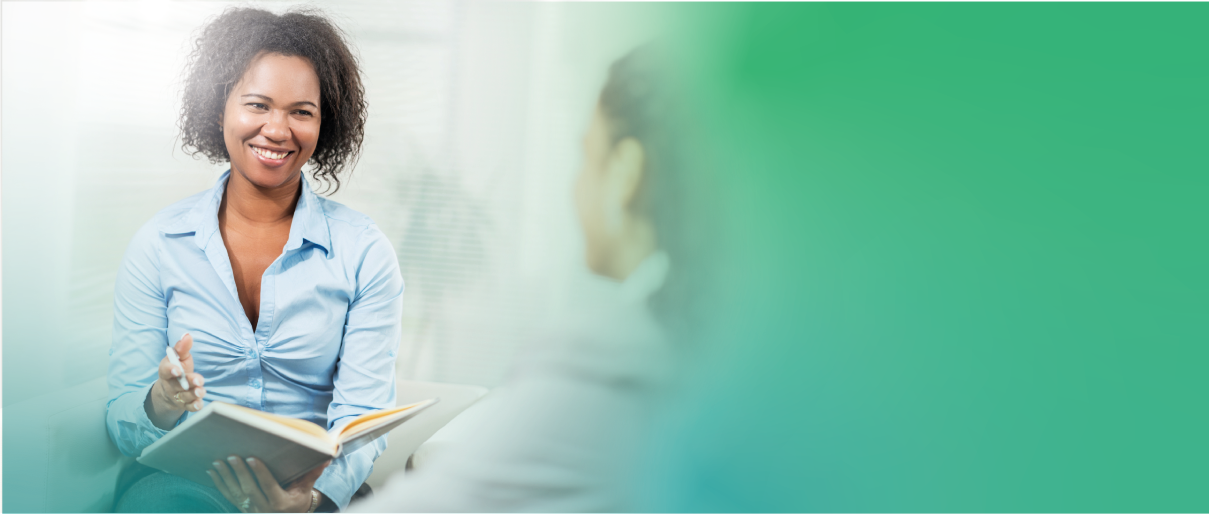 Una mujer le sonríe a otra que está en frente de ella. La cara de la segunda mujer no es visible en la imagen, y su cabeza está desenfocada y difuminada hacia verde. Las dos mujeres están conversando, y aunque el fondo está muy desenfocado para saber con certeza, parecen estar en una especie de oficina durante el día.